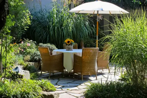Gedeckter Gartentisch mit Sonnenblumen unter einem hellen Sonnenschirm auf einer Natursteinterrasse, umgeben von hohen Gräsern und Stauden