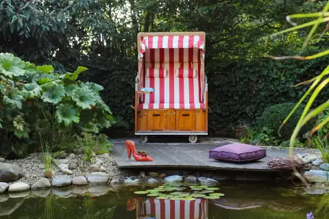 Hölzerner Strandkorb auf einem Holzdeck am Rand eines Gartenteichs mit Seerosen und üppigem Blattwerk