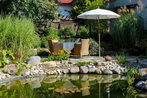 Gartenteich mit Sitzplatz unter einem Sonnenschirm, Findlingen und wassernahen Bepflanzungen
