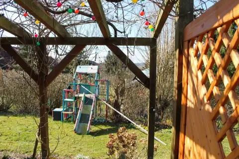 Ausgangssituation: Holzpergola mit Lichterkette, dahinter eine Rasenfläche mit Spielgerät